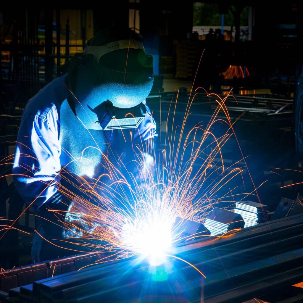 Welder sparks bright light while working on metal frame in a dark workshop.